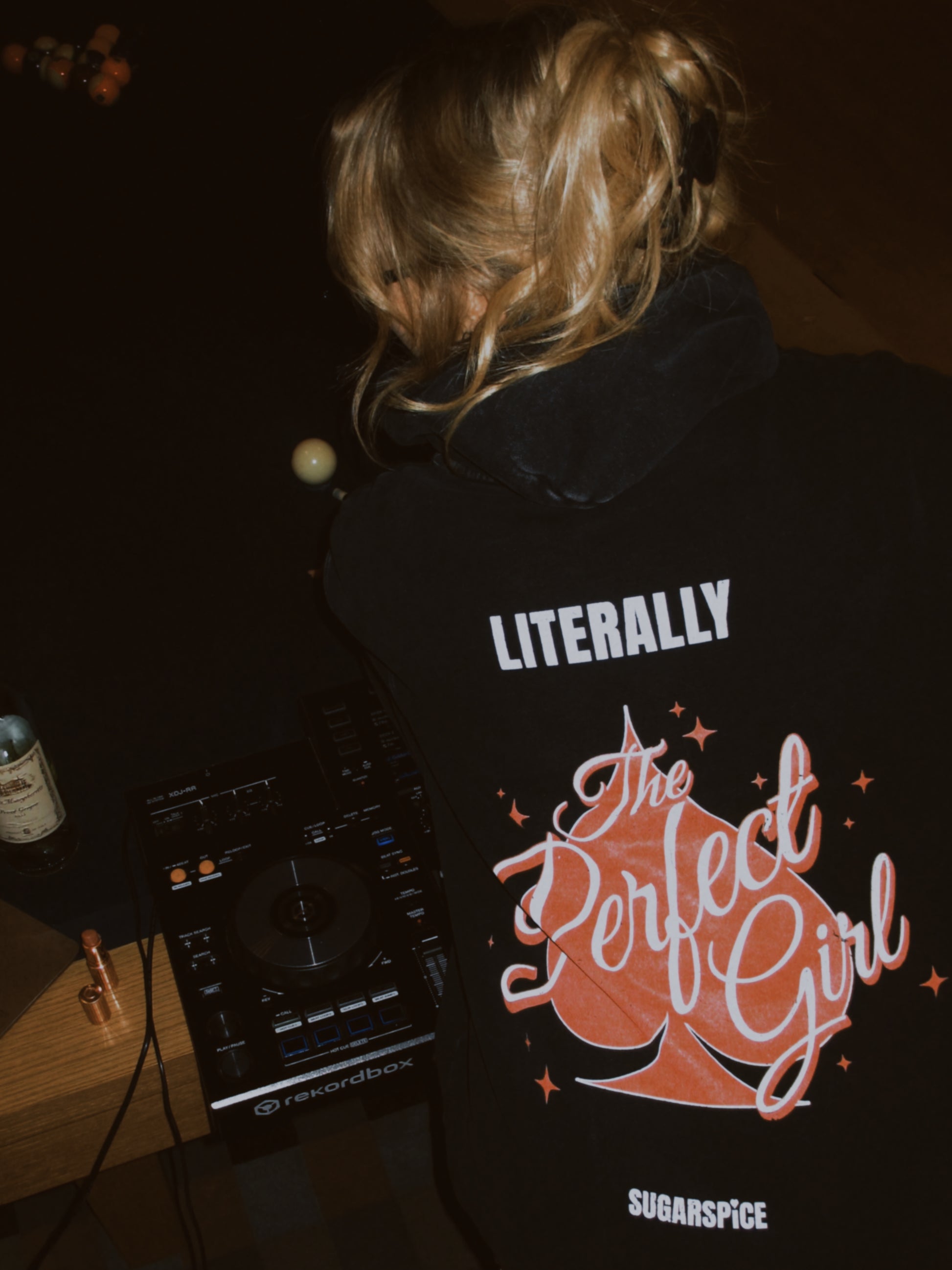 sugarspice wearing a black hoodie with text and graphics, standing in front of DJ equipment.